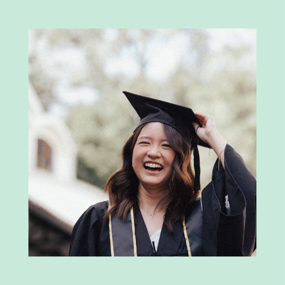 A young woman in a graduation cap and gown, smiling brightly as she holds her cap.