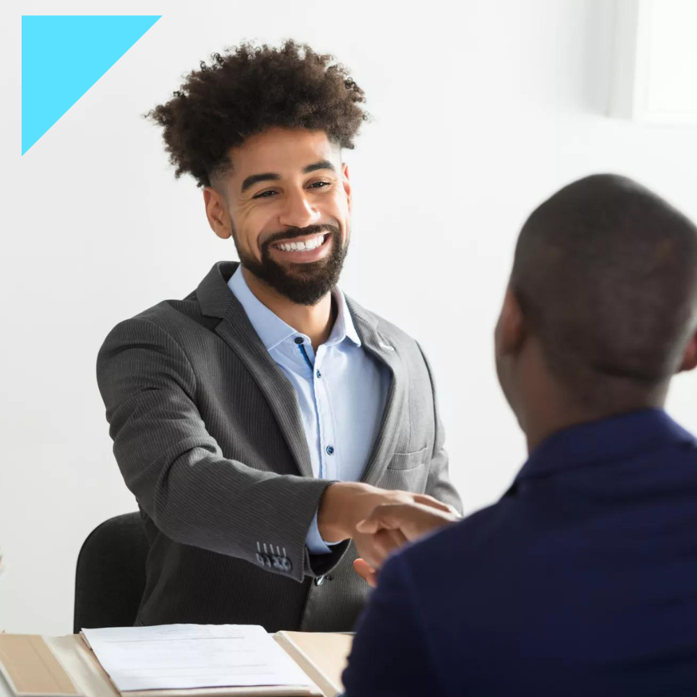 Smiling man in a suit shakes hands with another person across a desk during a job interview. A folder and documents are visible on the desk.