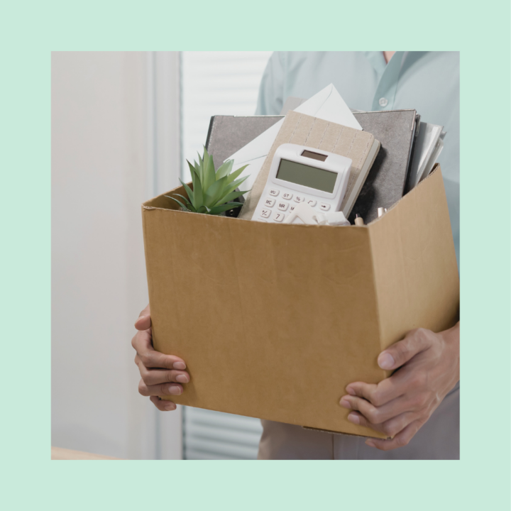 Person holding a cardboard box filled with personal belongings, including a plant, calculator, and documents, symbolizing being laid off.