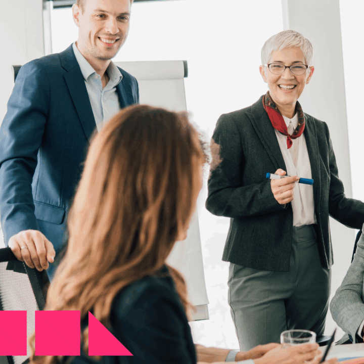 A multigenerational team of professionals in a modern office setting, with a younger man in a suit and an older woman with short gray hair leading a discussion by a flip chart. They are smiling and engaged, symbolizing collaboration and knowledge sharing between different generations in the workplace.