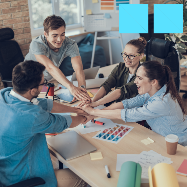 Four smiling coworkers stacking hands together over a collaborative workspace with papers, markers, and color swatches on the table