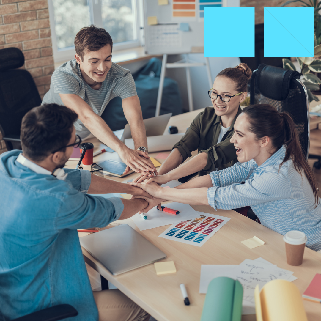 Four smiling coworkers stacking hands together over a collaborative workspace with papers, markers, and color swatches on the table