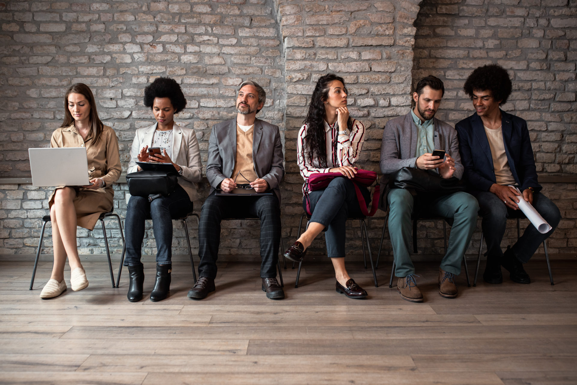 6 diverse professionals holding laptops, folders, and notepads sitting and waiting for their job interview
