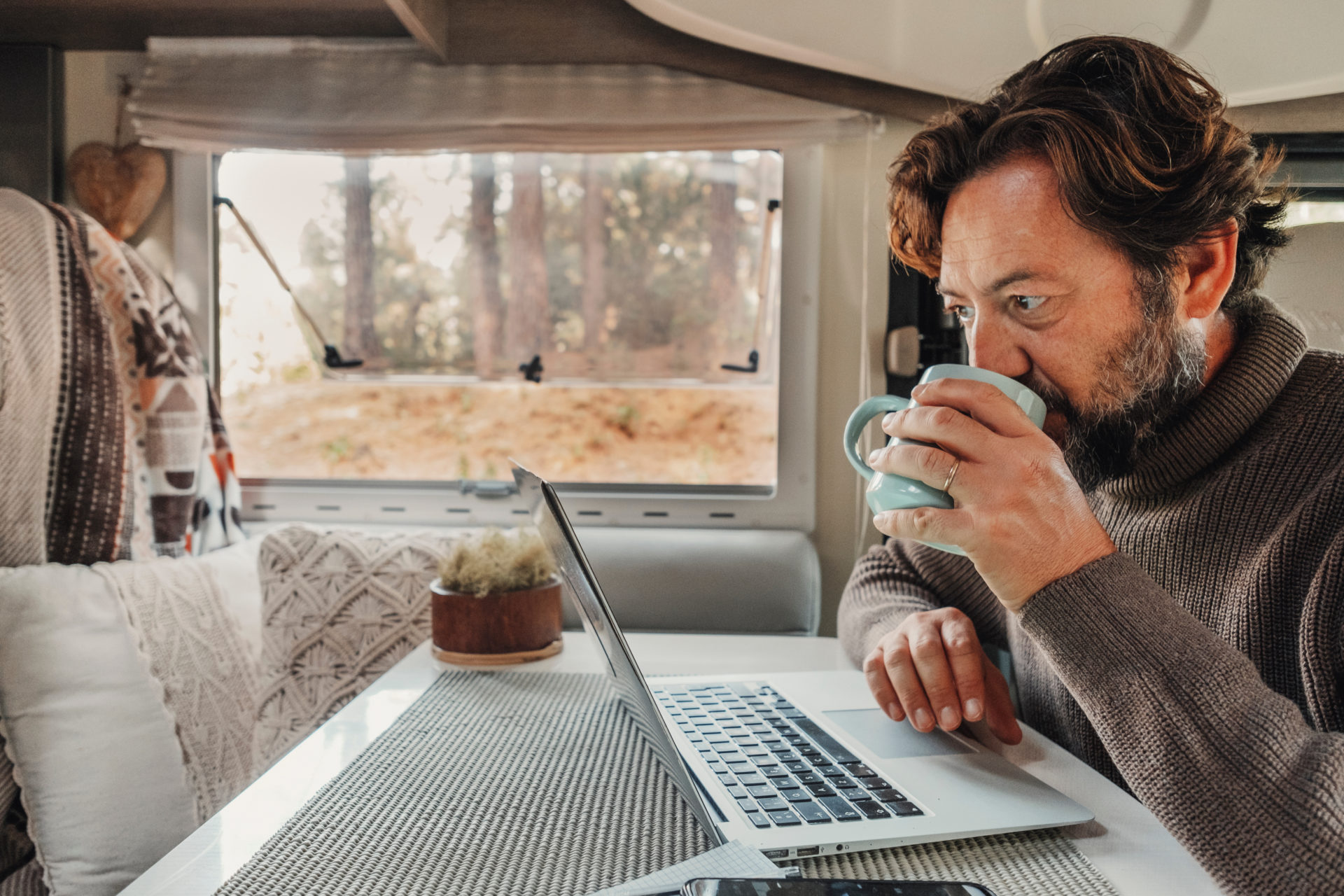 Man working on laptop computer inside a camper van with nature view outside the window.
