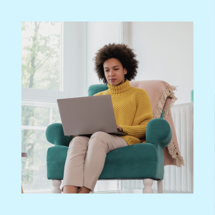 A young woman in a yellow sweater sits comfortably in a teal armchair, focused on her laptop. She appears to be reviewing or responding to an important message, possibly accepting a job offer. The setting is a cozy, well-lit living space with a blanket draped over the chair.