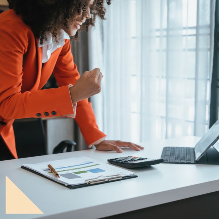 A professional woman in an orange blazer stands confidently at her desk, gesturing with determination while looking at her laptop. A clipboard with financial charts and a calculator are on the desk, suggesting she is preparing to ask for a raise or negotiate a salary increase.