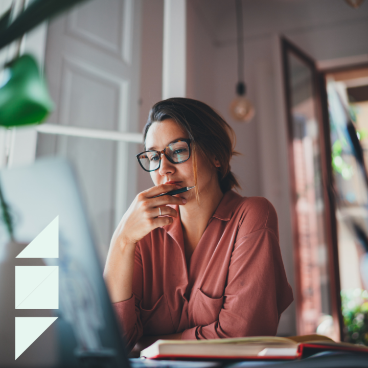 A thoughtful woman wearing glasses and a pink blouse sits at a desk, looking at her laptop screen while holding a pen to her chin. An open notebook lies beside her in a cozy, naturally lit workspace.