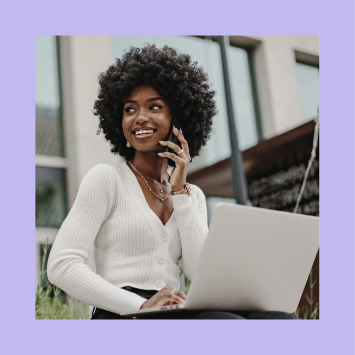 A young woman with curly hair sits outdoors with a laptop on her lap, smiling while talking on the phone. She wears a white ribbed cardigan and layered necklaces. The background features a modern building with large windows and greenery.