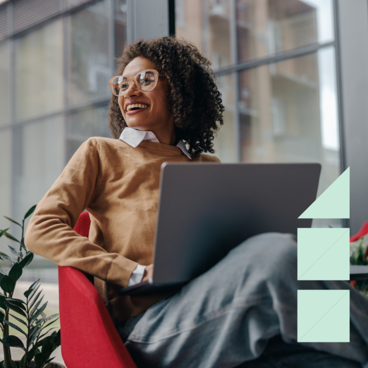 A smiling woman wearing glasses and a brown sweater sits in a red chair with a laptop on her lap. She looks off to the side in a modern, well-lit office space with large windows and greenery.