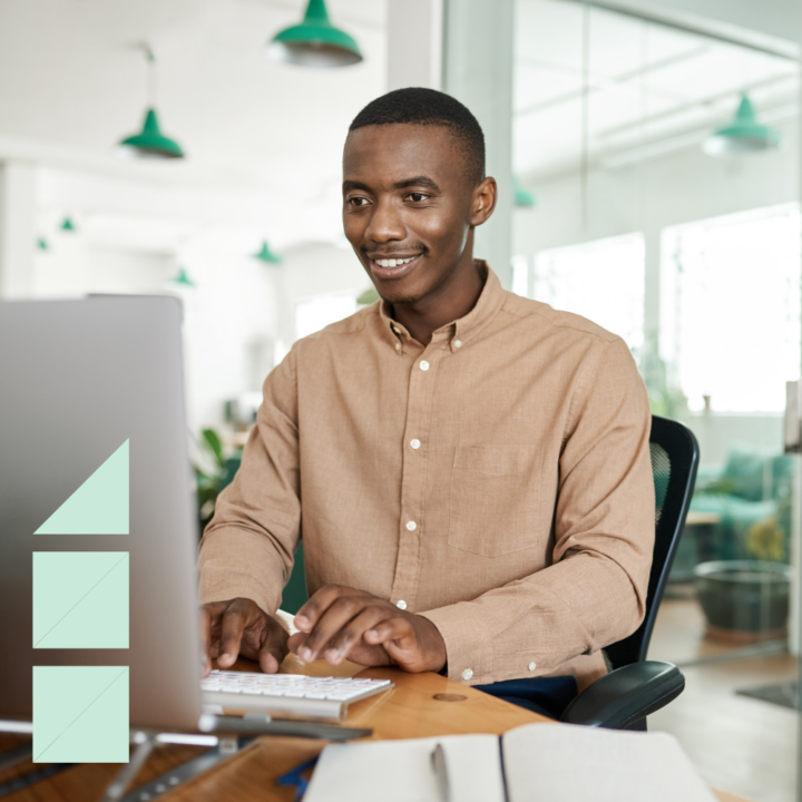 A young man in a beige button-up shirt smiles while working on his computer in a modern, well-lit office. He sits at a wooden desk with an open notebook and pen. The background features glass partitions, green pendant lights, and office plants.
