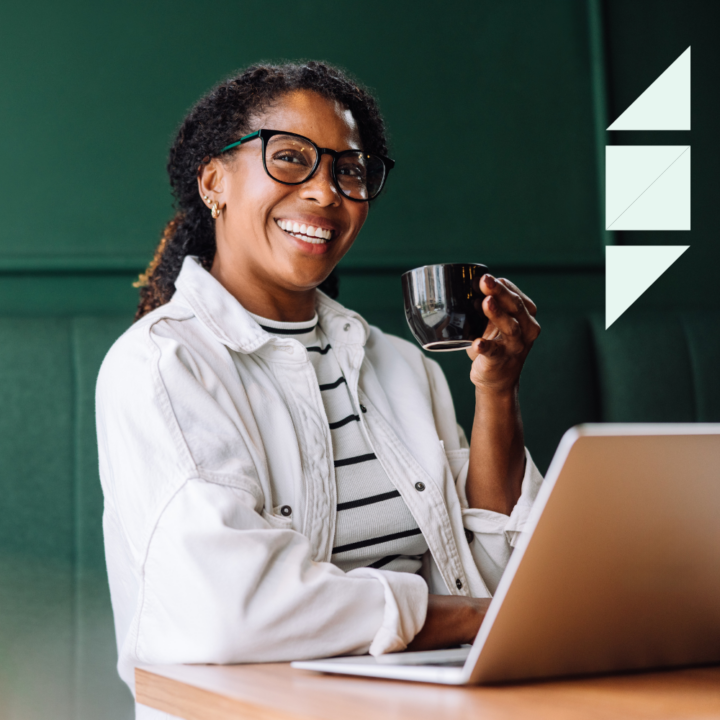 A smiling woman wearing glasses and a white jacket sits at a café table with a laptop, holding a black coffee cup. She appears relaxed and engaged in her work.