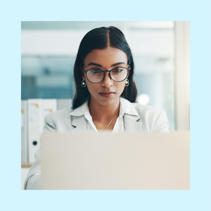 A professional woman in a light-colored blazer and glasses is focused on her laptop in an office setting. With a determined expression, she appears to be working on an important document, drafting a promotion request.