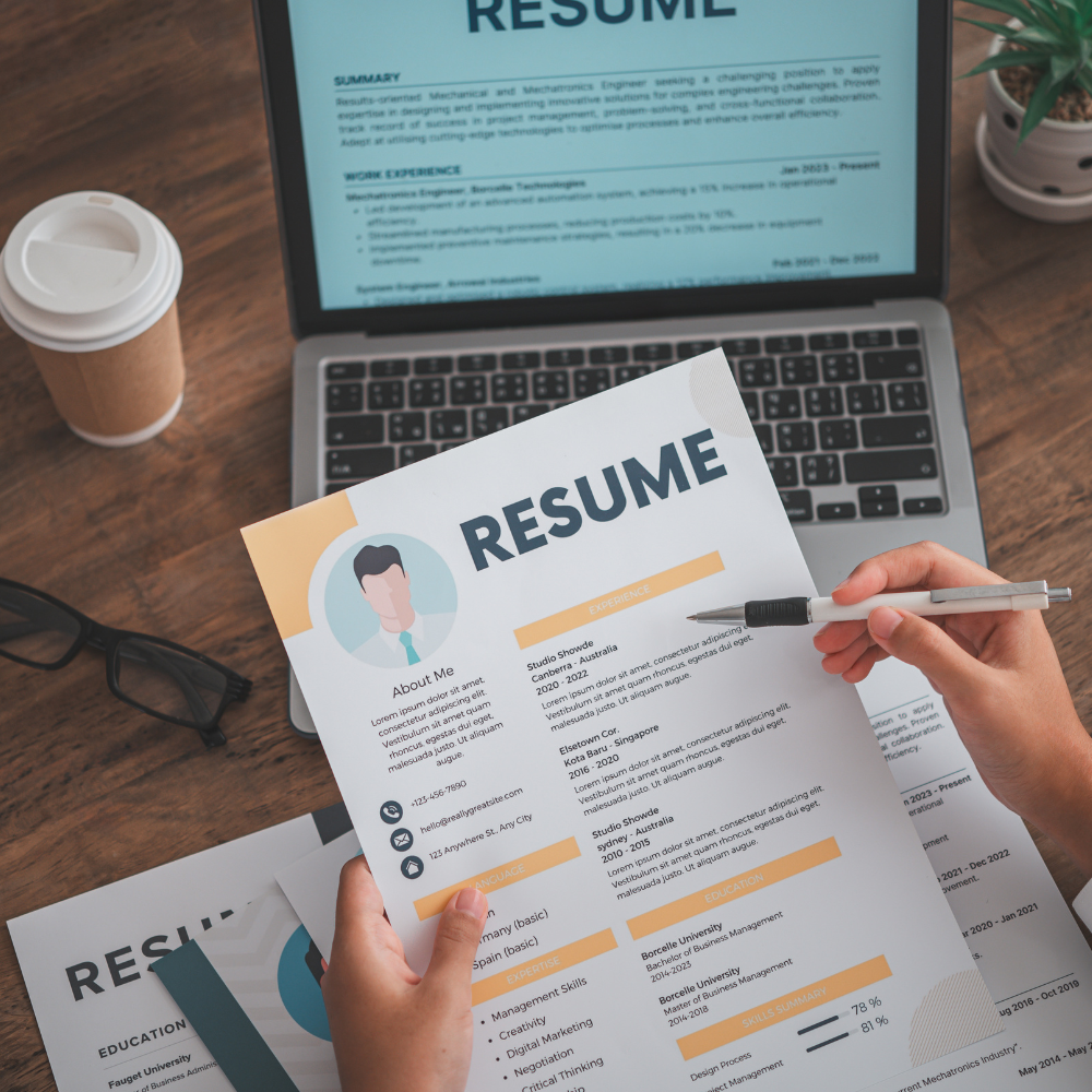 A person reviewing a visually appealing, creative resume with a modern design, featuring colorful sections and a profile illustration. A laptop displaying a digital version of the resume, along with a coffee cup, glasses, and a plant, are placed on a wooden desk, creating a professional yet artistic workspace.
