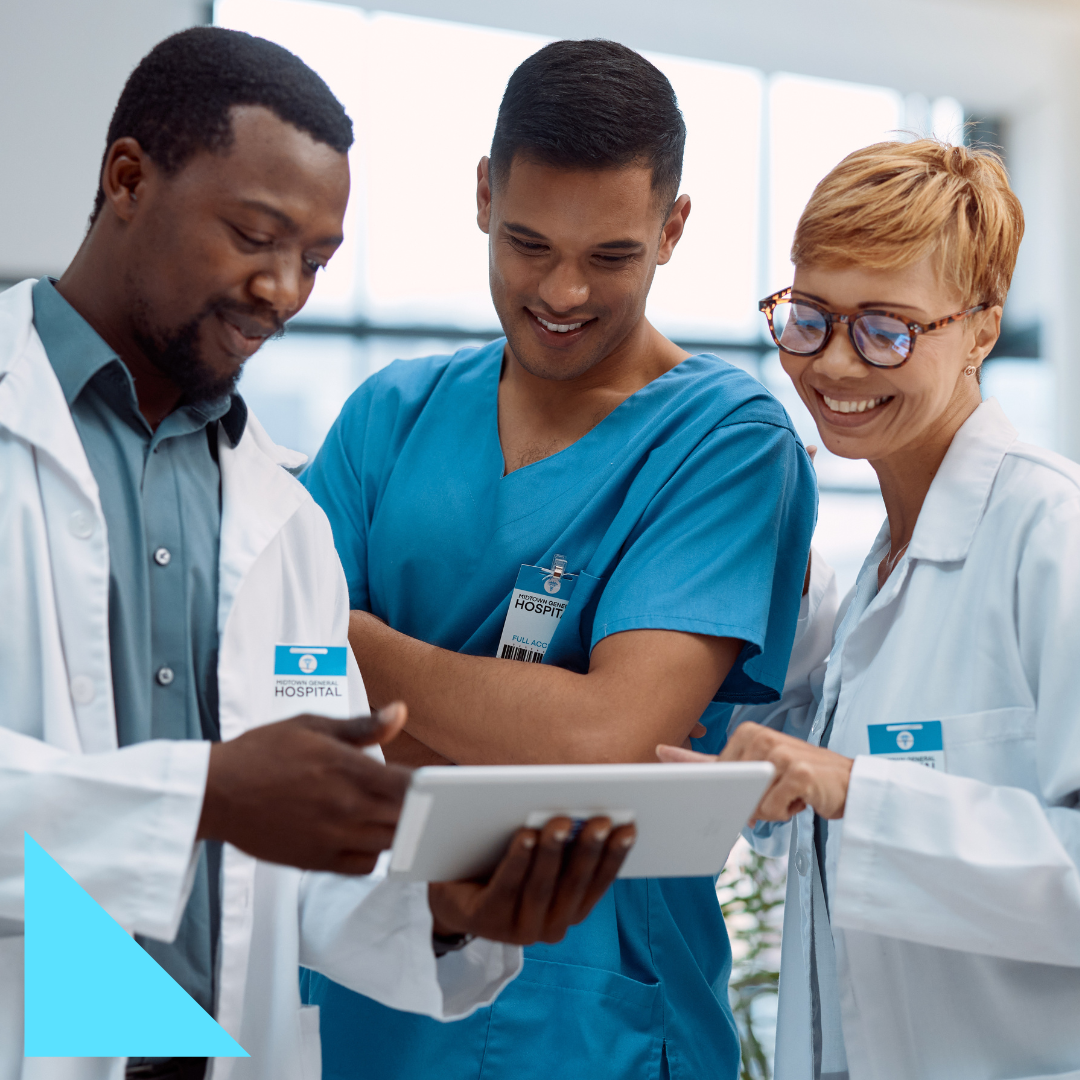Three healthcare professionals—a doctor in a white coat, a nurse in blue scrubs, and a clinician with glasses—review information together on a tablet in a hospital setting, smiling while collaborating on patient care and clinical workflows.