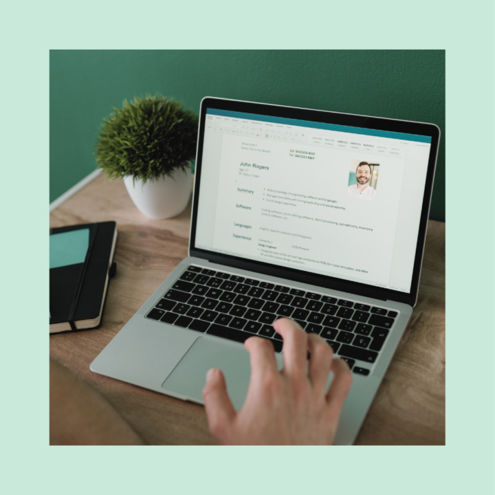 A job seeker reviewing a CV on a laptop screen, featuring a professional headshot, summary, skills, and experience sections. The workspace includes a notebook and a potted plant, set against a green wall.