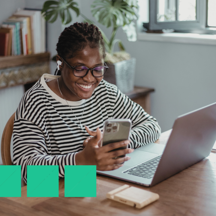 A young woman wearing glasses and a striped shirt smiles while looking at her phone, with a laptop open on the table beside her. She is wearing earbuds and appears engaged in a conversation, possibly negotiating a job offer remotely. The setting is a cozy home workspace with books and plants in the background.