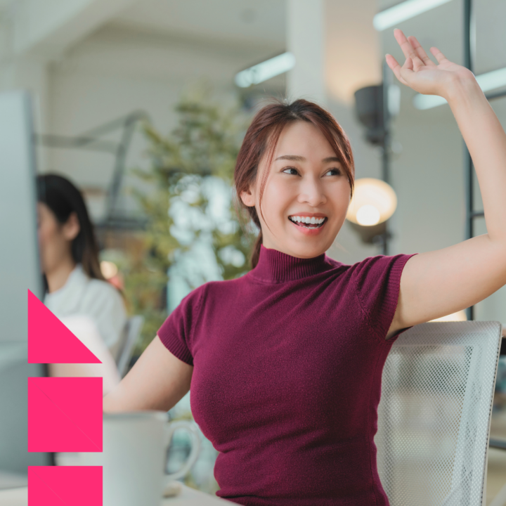 A cheerful woman in a burgundy top sitting at her desk in a modern office, smiling and waving as she looks toward someone off-camera. The environment is bright and welcoming, with a colleague working in the background.