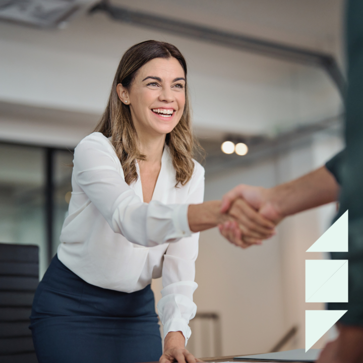 A professional woman in a white blouse and navy skirt smiles as she shakes hands with an interviewer. She leans slightly forward in a modern office setting