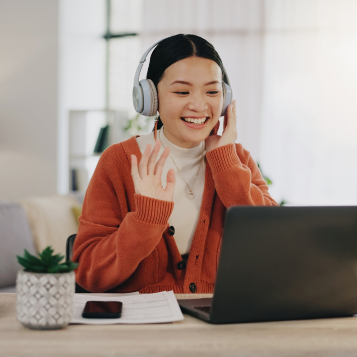 A young woman wearing headphones and an orange cardigan smiles and waves during a virtual meeting on her laptop. She sits at a desk with documents, a smartphone, and a small potted plant.