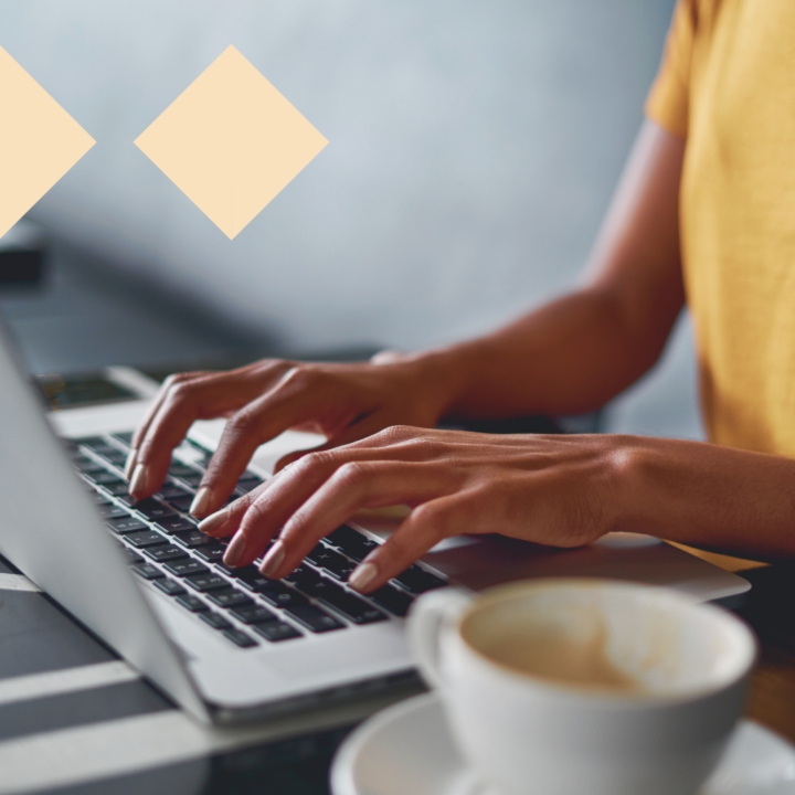 Close-up of a person in a yellow shirt typing on a laptop at a café table. A cup of coffee sits nearby, creating a cozy and focused work environment.