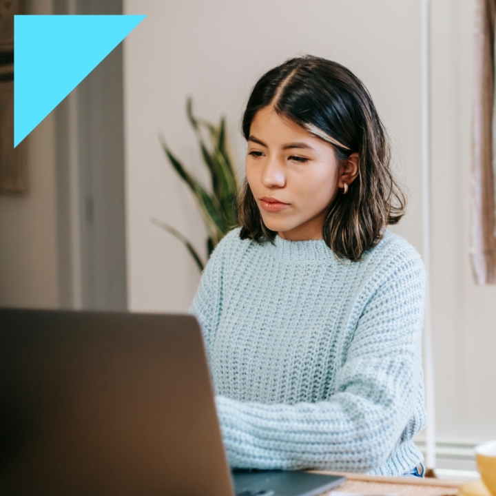 A young woman in a light blue sweater sits at a desk, focused on her laptop with a serious expression. She appears to be carefully composing a message, possibly declining a job offer.