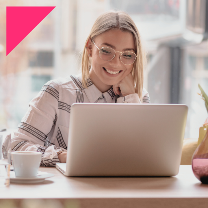 A smiling woman with glasses and a plaid shirt sits at a café table, working on her laptop. A cup of coffee and decorative vase are nearby, creating a warm and inviting atmosphere.