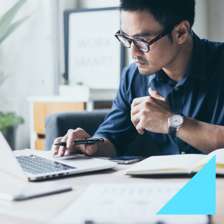 A focused man wearing glasses and a navy blue shirt works on his laptop while holding a pen and smartphone. He sits at a desk with an open notebook and resignation letter. The background features a modern office.