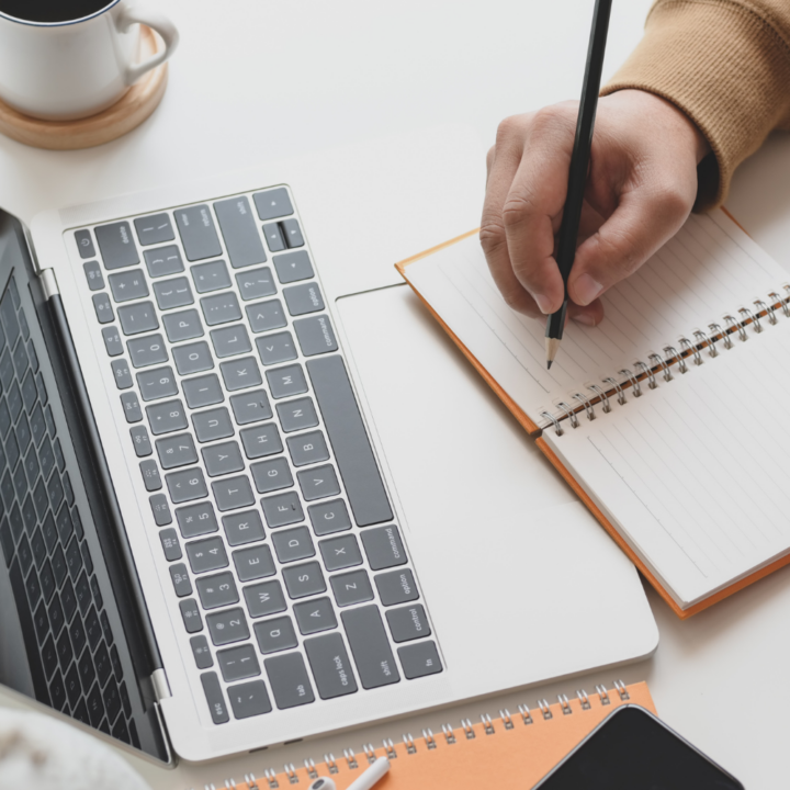 A person writing in a spiral notebook next to an open laptop, preparing a student resume. The workspace includes a cup of coffee, a smartphone, and additional notebooks, creating a focused and organized setting.