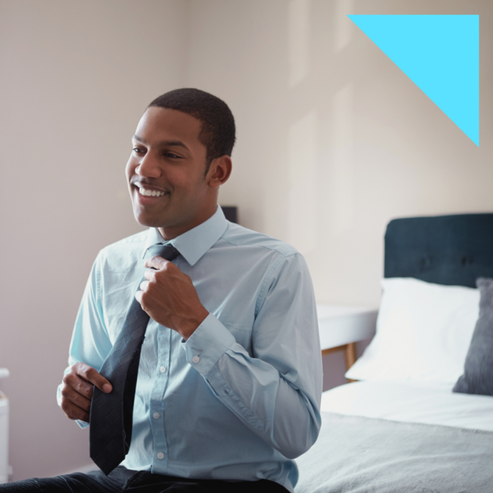 A smiling man in a light blue dress shirt sits on the edge of a bed while adjusting his tie, preparing for his first day of work. The room is well-lit with a neatly made bed