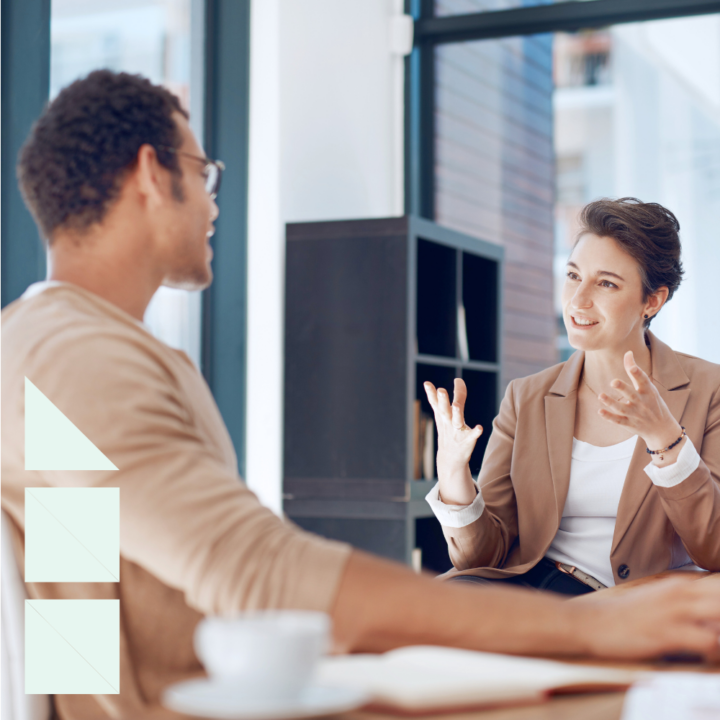 A professional woman in a brown blazer engaged in a transfer conversation with her male boss in a modern office setting. She is gesturing expressively while speaking, and the man opposite her listens attentively with a coffee cup and notebook in front of him.