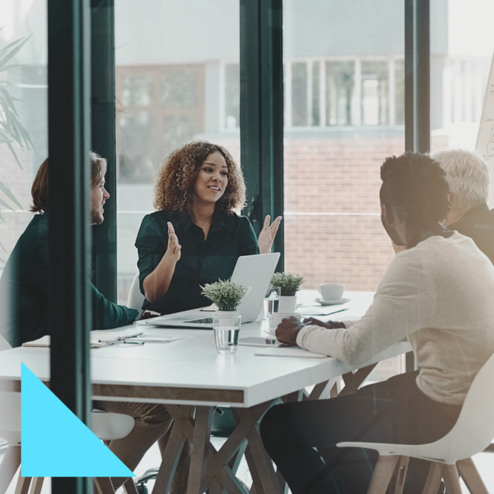 A diverse group of four professionals is engaged in a business meeting in a modern office with large windows. A woman with curly hair, wearing a dark blouse, is speaking and gesturing with her hands while others attentively listen. A laptop, notebooks, and glasses of water are on the white table. The setting is bright, with natural light streaming in through the glass walls.