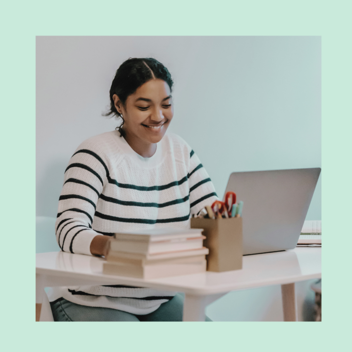 A young woman in a striped sweater sits at a white desk, smiling as she works on her laptop. A stack of books and a container filled with pens and pencils are nearby, suggesting she is writing an internship cover letter.