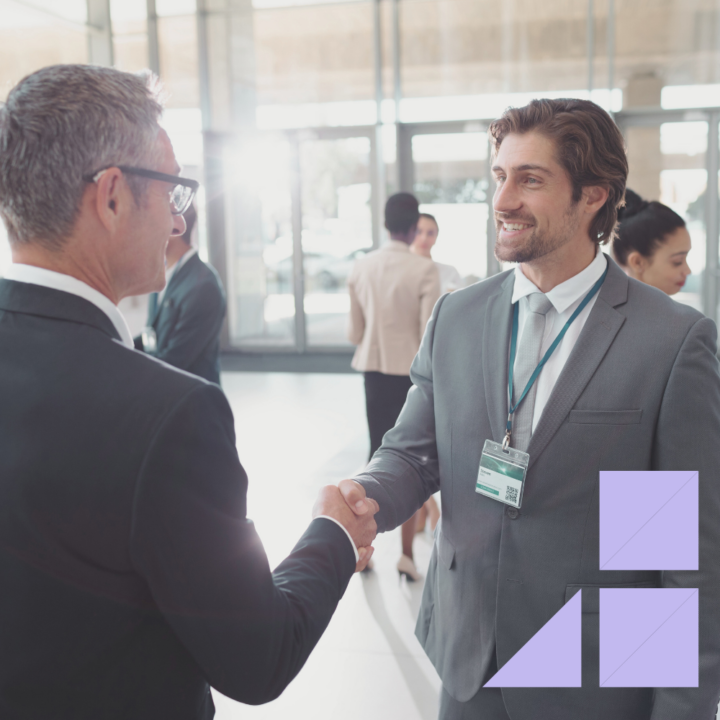 Two professionally dressed men shaking hands at a networking event in a modern conference hall. The man in the foreground wears a suit and ID badge, smiling as they engage in conversation, with other attendees mingling in the background.