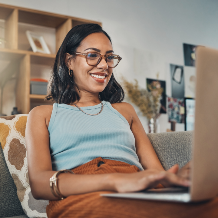 A smiling woman wearing glasses and a blue sleeveless top sits on a cozy couch, typing letter of intent on her laptop. She is in a stylish, well-decorated living space with shelves and wall art in the background.
