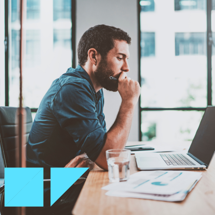 A thoughtful man in a dark blue shirt sits at a modern office desk, looking at his laptop screen while holding a pen to his chin. Documents with charts and a glass of water are on the desk, with large windows providing natural light in the background.