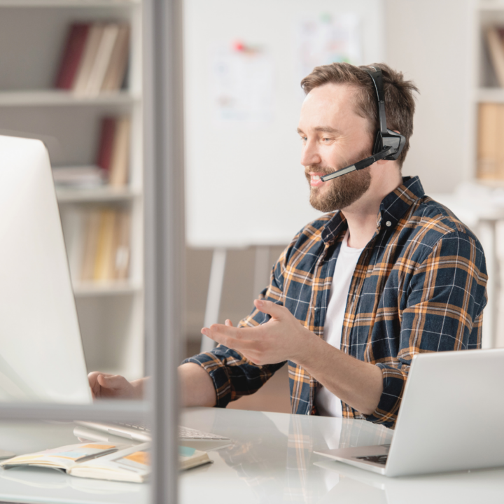A man wearing a plaid shirt and a headset sits at a desk, engaged in a video call on his computer. He gestures with his hand while speaking, appearing to communicate professionally offering an apology to a client.