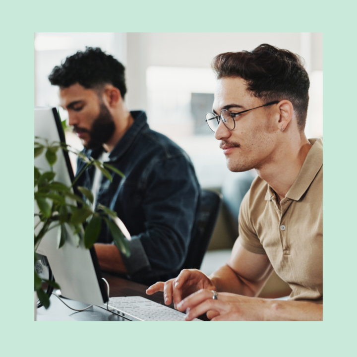 Two young professionals working at desktop computers in a modern office setting. The man in the foreground, wearing glasses and a beige polo shirt, is focused on his screen, while another colleague in a denim jacket is working in the background.
