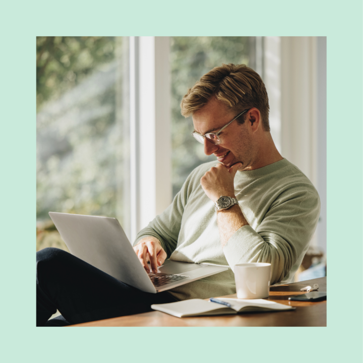 A smiling man wearing glasses and a light green sweater sits at a wooden desk, working on his laptop. A cup of coffee, notebook, and pen are nearby, with natural light streaming through large windows.