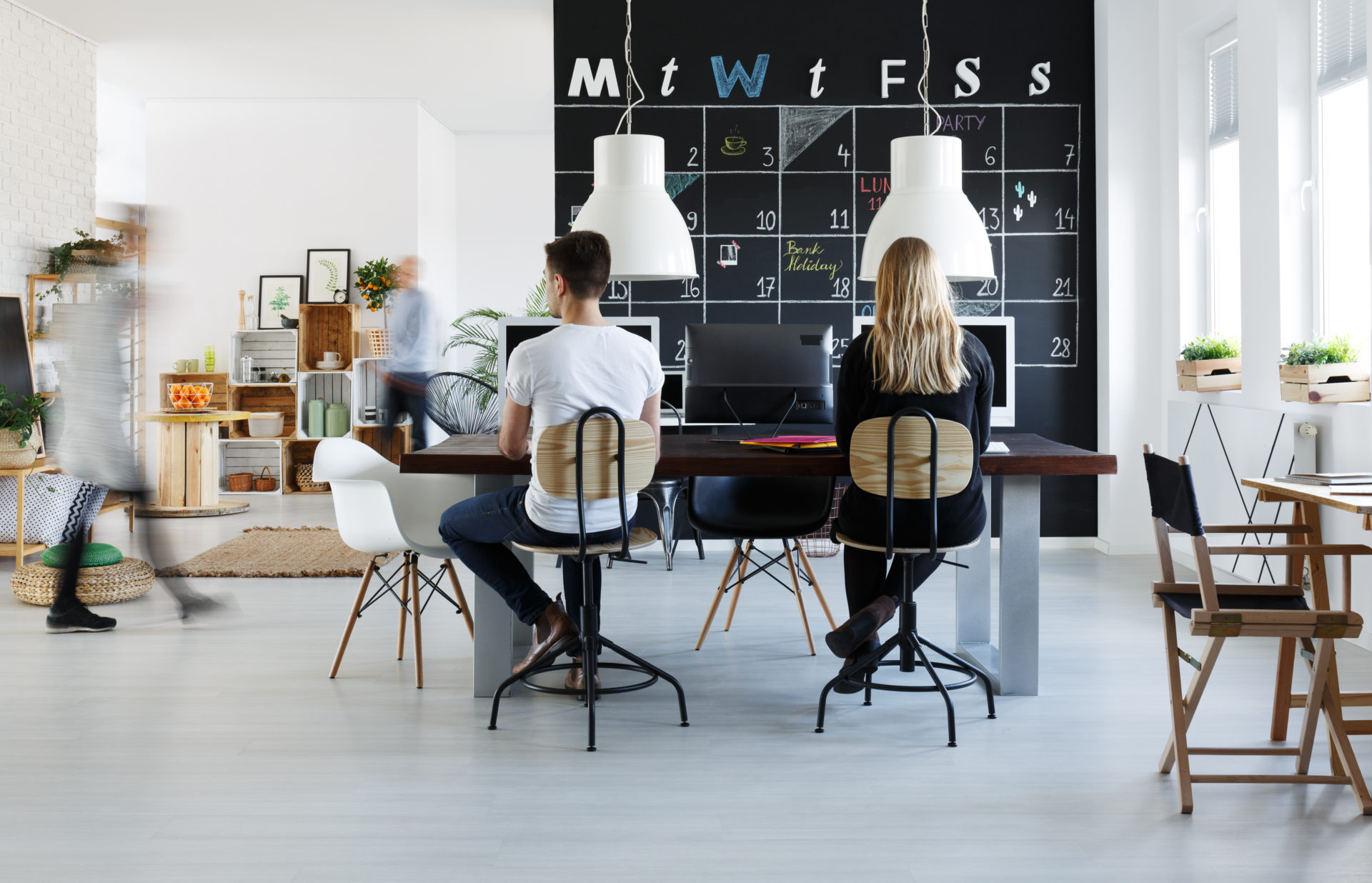 Office space with chalkboard calendar on the wall. Desk center with male and female professional looking at their computer during their 4 day workweek.