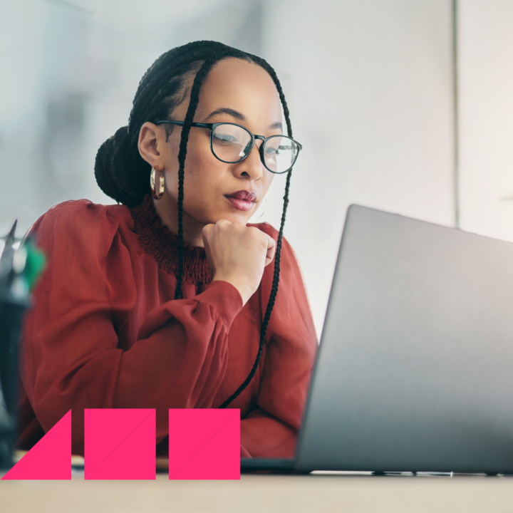 A professional woman wearing glasses and a red blouse sits at a desk, intently focused on her laptop screen. Her hand rests under her chin as she thoughtfully composes an email in a well-lit office setting.
