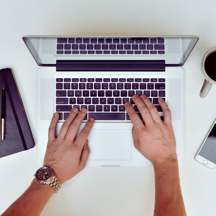 Top-down view of a person's hands typing on a laptop keyboard. A notebook, pen, coffee cup, and smartphone are neatly arranged on the desk, creating a productive workspace.