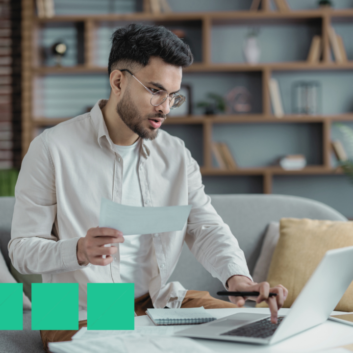A focused man wearing glasses and a beige shirt sits on a couch, reviewing a personal leave document while working on his laptop. A notebook and papers are spread out in front of him in a cozy home office setting with a modern bookshelf in the background.