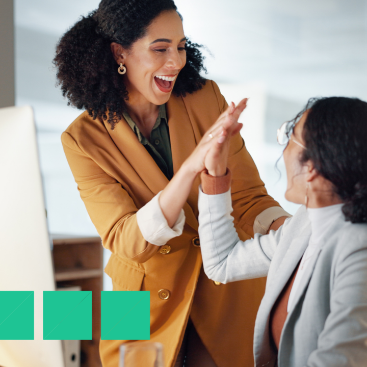 Two professional women in a modern office celebrating a work achievement with a high-five. One is wearing a mustard blazer and smiling excitedly, while the other, in a light-colored blazer, responds with enthusiasm.