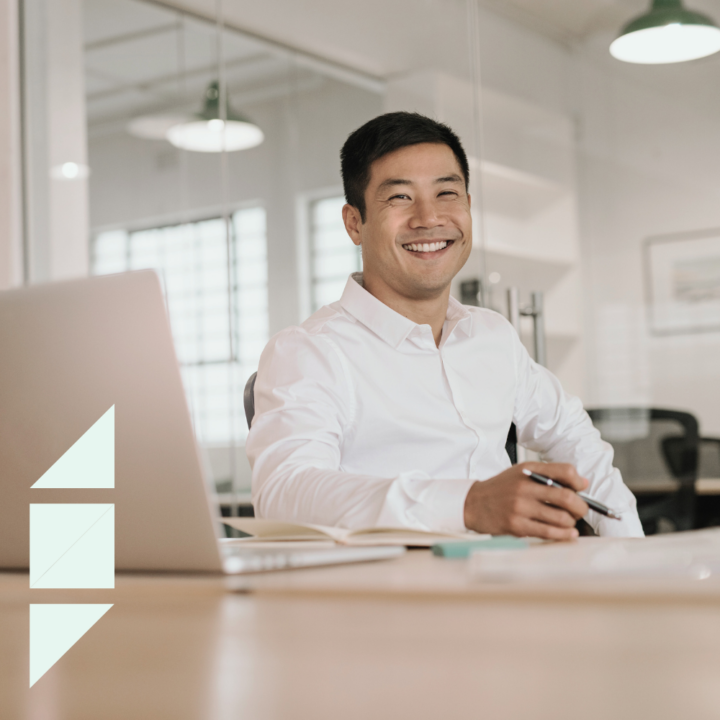 smiling man in a white shirt sits at a desk in a modern office, holding a pen and appearing to work on a document. A laptop is open beside him, suggesting he is engaged in writing or planning, working on a personal statement.