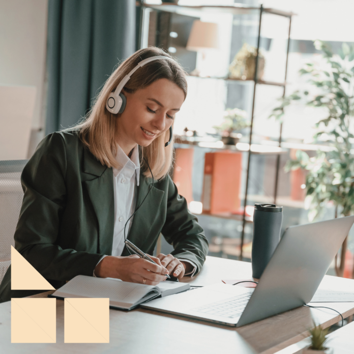 A young woman with blonde hair wears a headset while taking notes in a notebook during a virtual meeting. She is seated at a desk with a laptop, a reusable water bottle, and office supplies. The background features a well-lit, modern office with shelves, plants, and large windows.