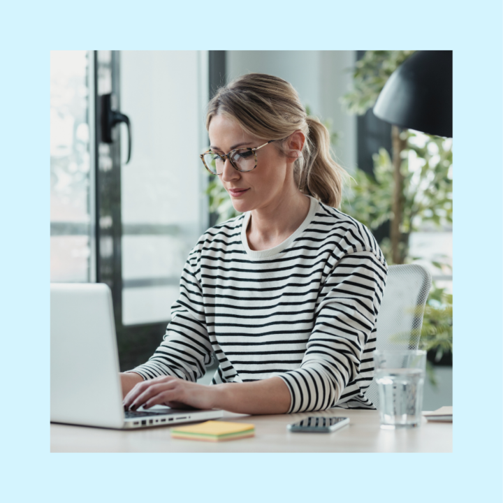 A woman wearing glasses and a striped sweater works on her laptop in a bright, modern office. She is seated at a desk with a notebook, smartphone, and a glass of water.