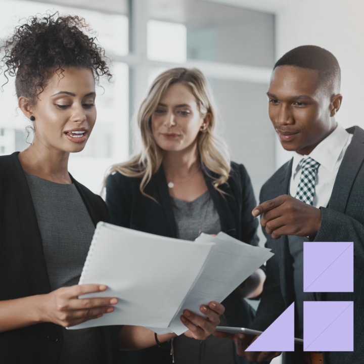Three professionally dressed colleagues reviewing a business proposal together in a modern office setting. One woman holds a document while the others look on and discuss the details.
