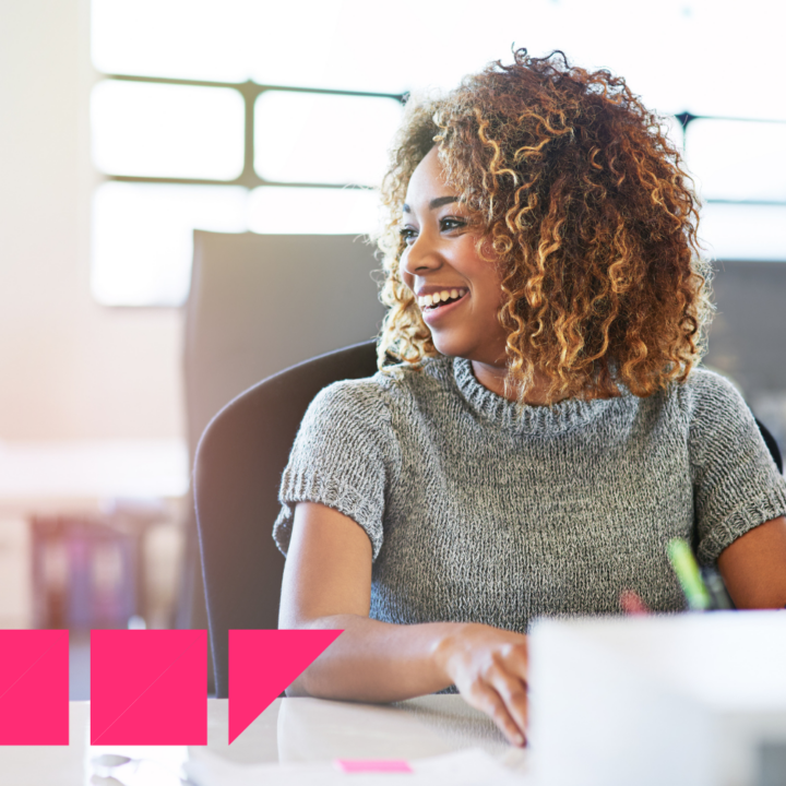 A young woman with curly hair smiles while sitting at a desk in a bright, modern office. She wears a gray knitted top and appears engaged in a conversation. Papers, laptop, pens, and office supplies are on the desk.