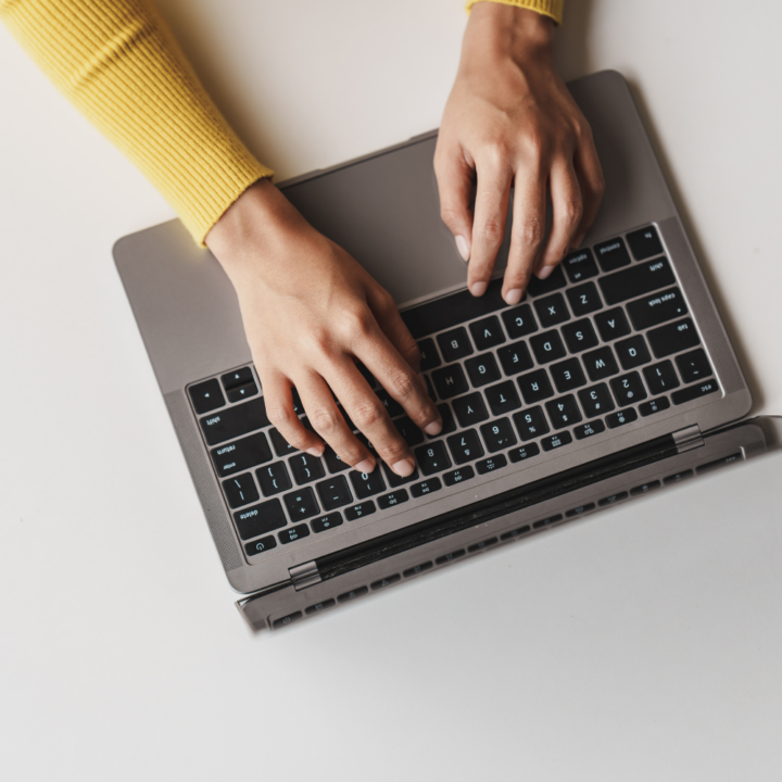 Close-up of a person's hands typing on a laptop keyboard. The individual is wearing a yellow sweater, and the workspace has a clean, minimalistic white background.