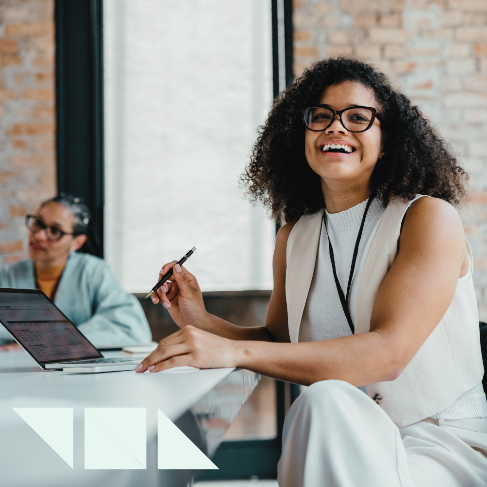 A smiling woman recruiter wearing glasses and business attire sits at a desk with a new tech software on a laptop and pen in hand, appearing engaged and confident in a modern office setting. Another woman is seated in the background.